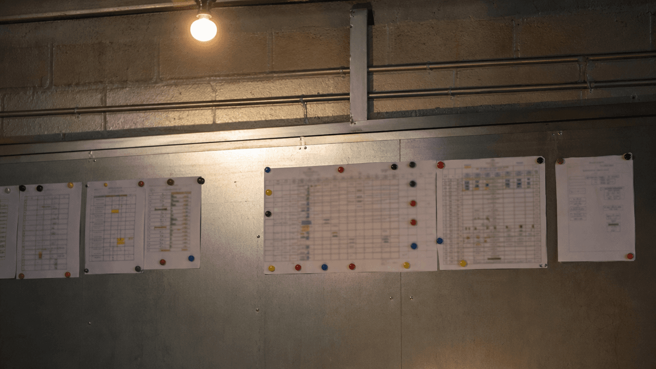 Plannings papier punaisés sur le mur d'un local technique, éclairés par une ampoule nue